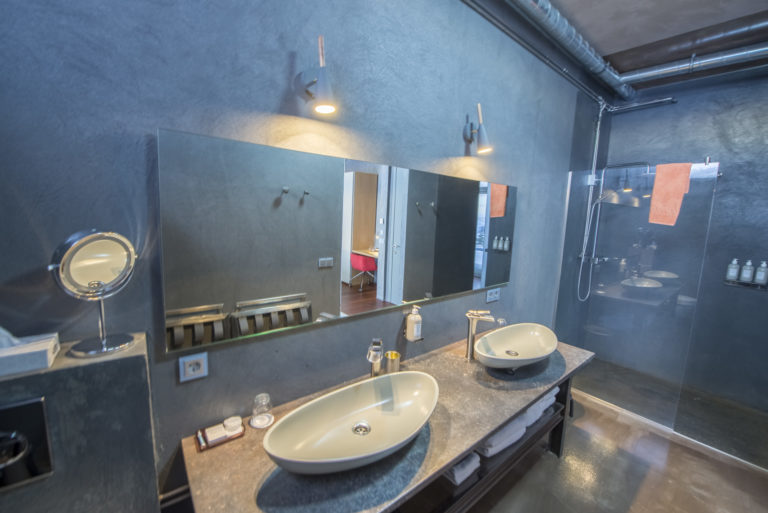 A modern bathroom with a dark color scheme. The walls and floor are finished in a slate gray texture, and the ceiling ductwork is exposed, painted black. On the right, a clear glass shower stall reveals a matching interior with a white basin and bright coral towels hanging inside. The vanity area features a large mirror reflecting the room, two oval white sinks atop a granite countertop, and a smaller magnifying mirror. There are minimalistic wall-mounted faucets and a selection of toiletries arranged neatly on the counter. The room is well-lit with wall-mounted lights on either side of the mirror casting a warm glow.