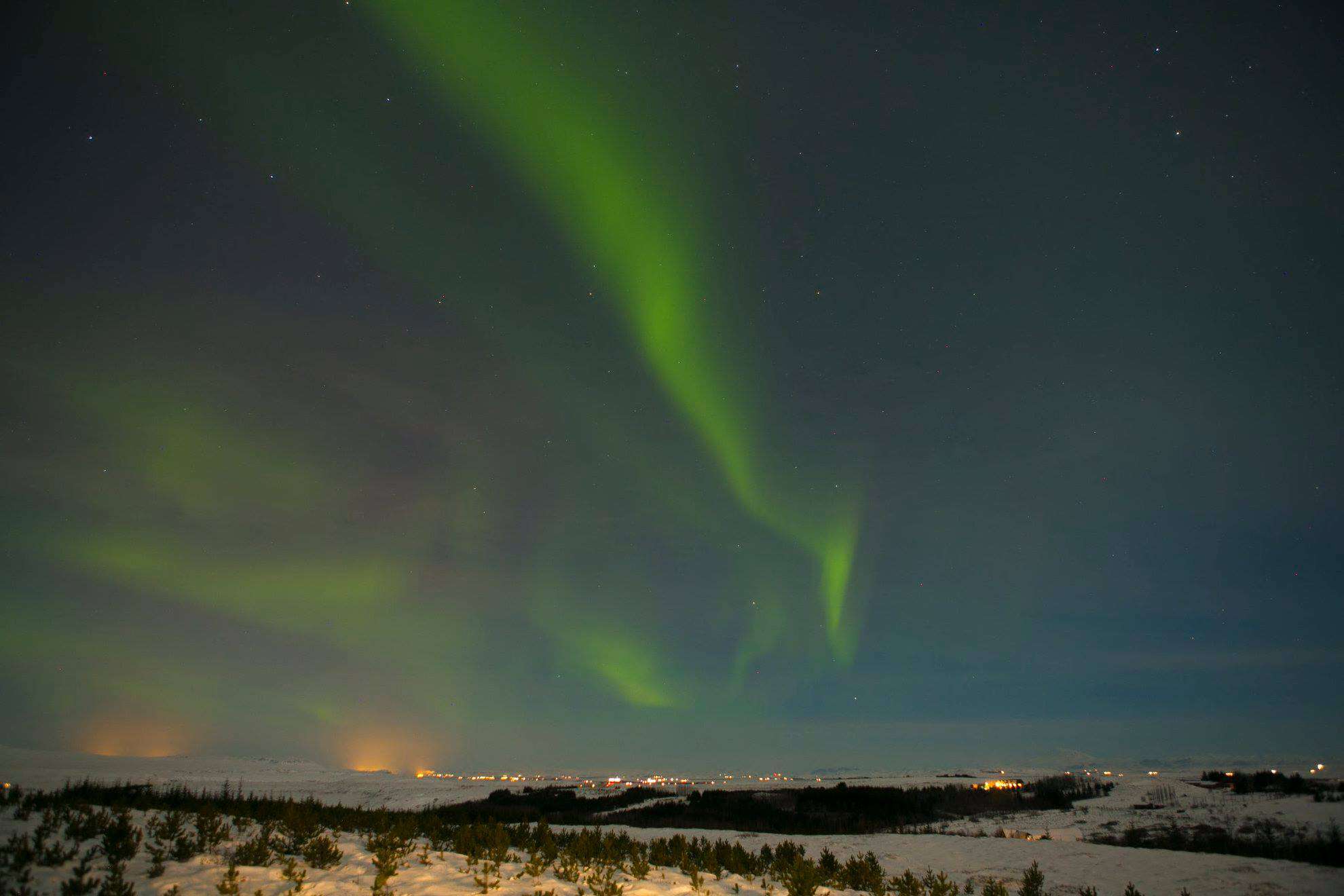  The Northern Lights (Aurora Borealis) illuminate the night sky with a vibrant display of green ribbons above a snowy landscape. The scene is dotted with small lights from a distant settlement, and the stars are visible in the dark sky above.