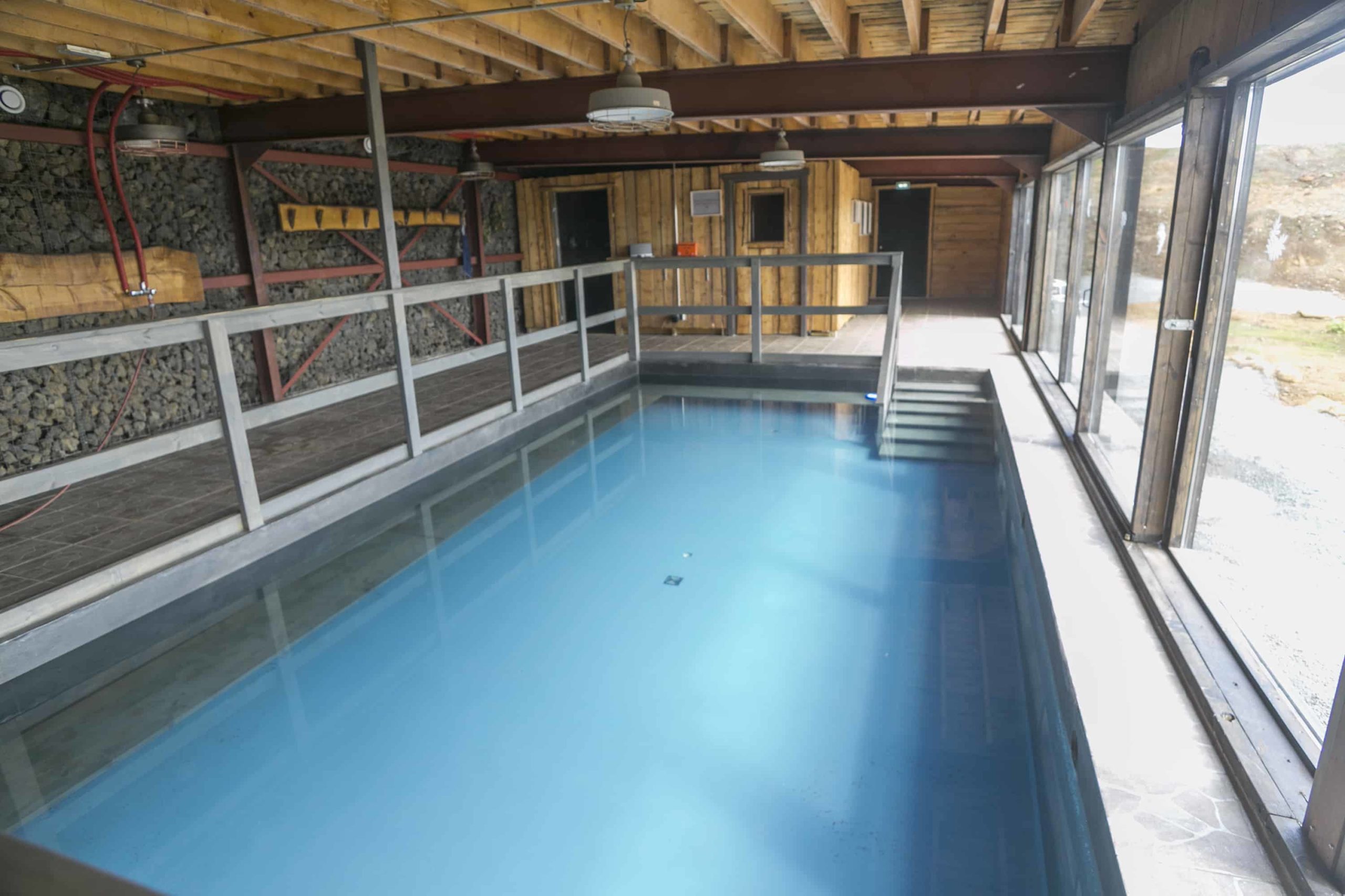 An indoor swimming pool within a rustic setting, featuring exposed wooden beams and stone walls. The pool is calm, reflecting the wood-paneled ceiling, and is flanked by a metal railing. Large windows allow natural light to enter, and a wooden sauna box is visible at the far end of the room
